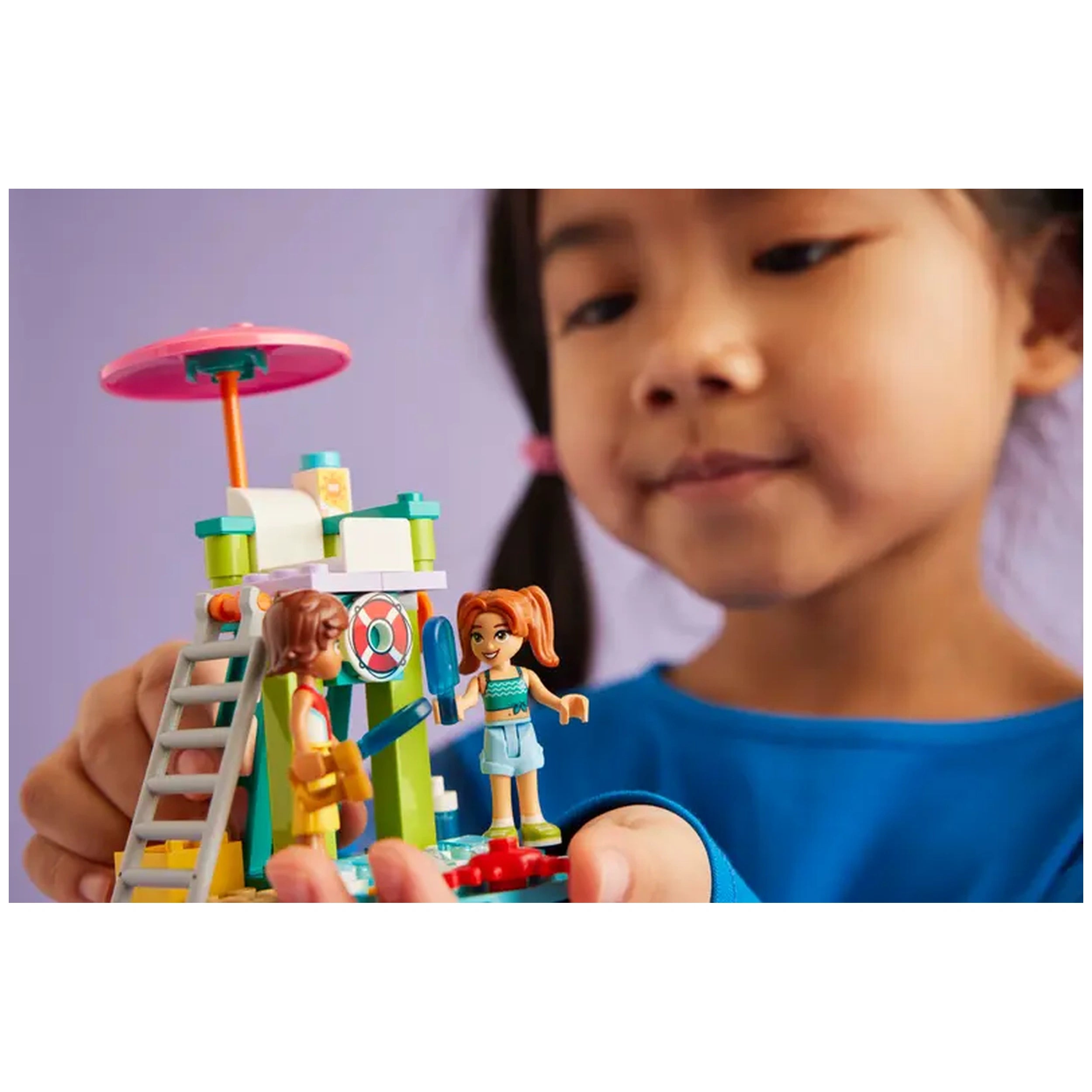 A child holds a colorful LEGO Friends Beach Water Scooter set featuring two mini-dolls and a lifeguard tower, set against a purple background.