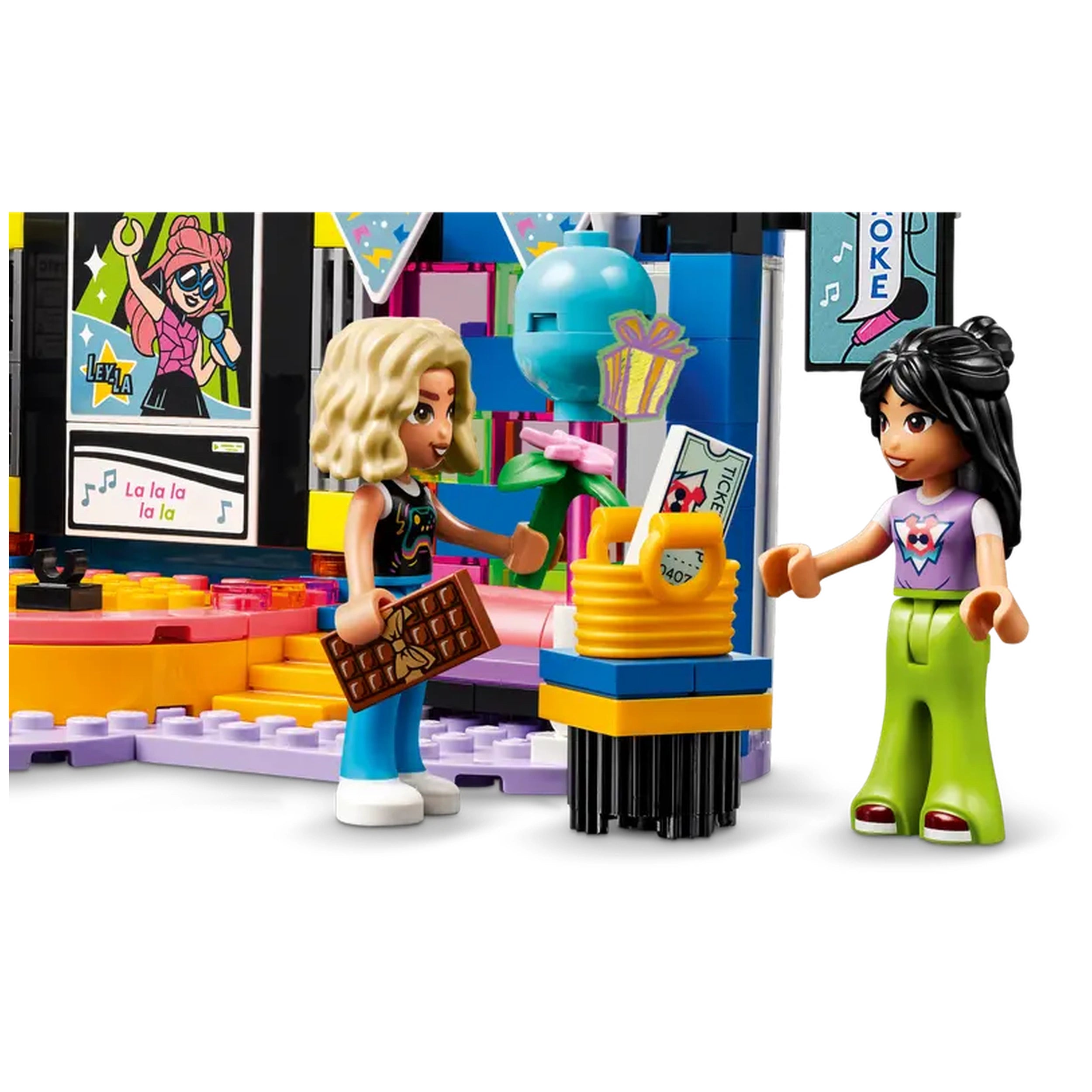 Two LEGO Friends characters at a karaoke party, one holding a chocolate bar and a flower, the other with a microphone. Colorful stage elements in the background with music-themed decorations and a ticket display.