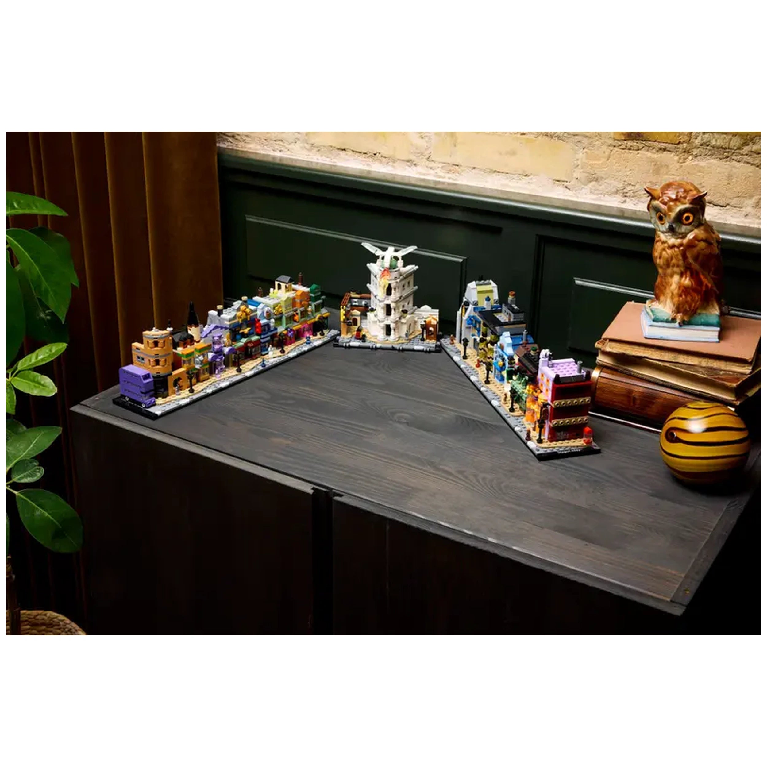 Colorful LEGO® Harry Potter™ Diagon Alley™ microscale model displaying iconic shops on a dark wooden surface, with a decorative owl and potted plant in the background.