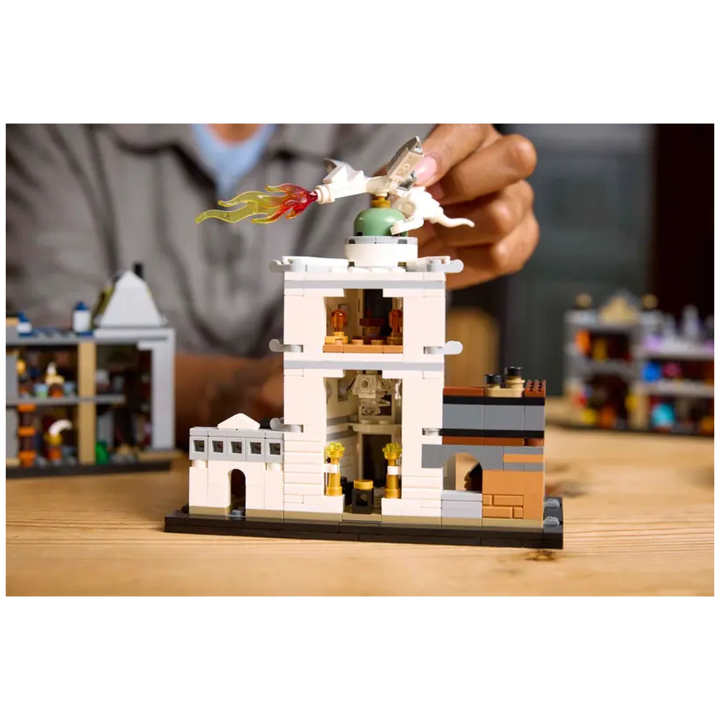 A person’s hand reaches for a LEGO® structure featuring the iconic Ollivanders™ wand shop, with colorful details and flames atop the roof. A second building with visible interior scenes is set on a wooden surface.