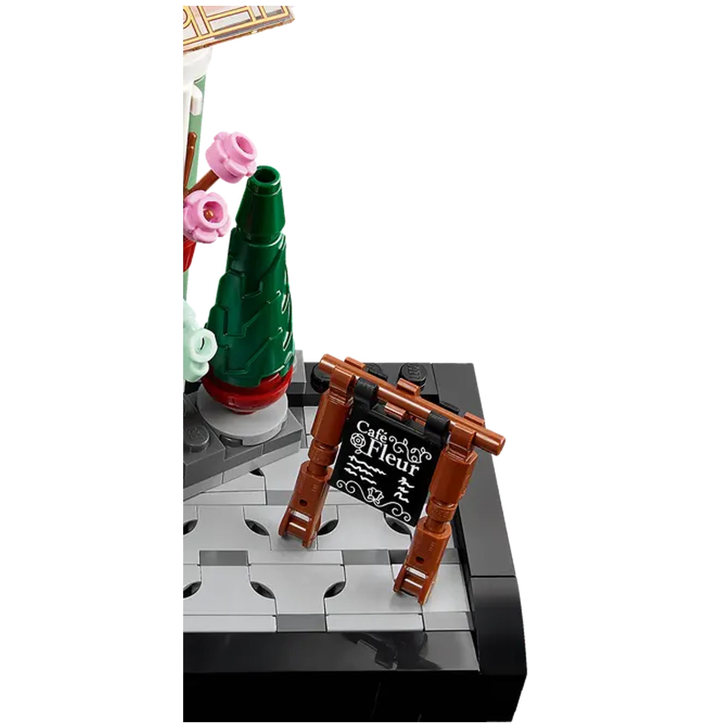 A small brown wooden sign with black chalkboard detailing reads "Café Fleur," next to a green coniferous plant and pink flowers. The scene is set on a gray brick sidewalk, capturing a charming café atmosphere.