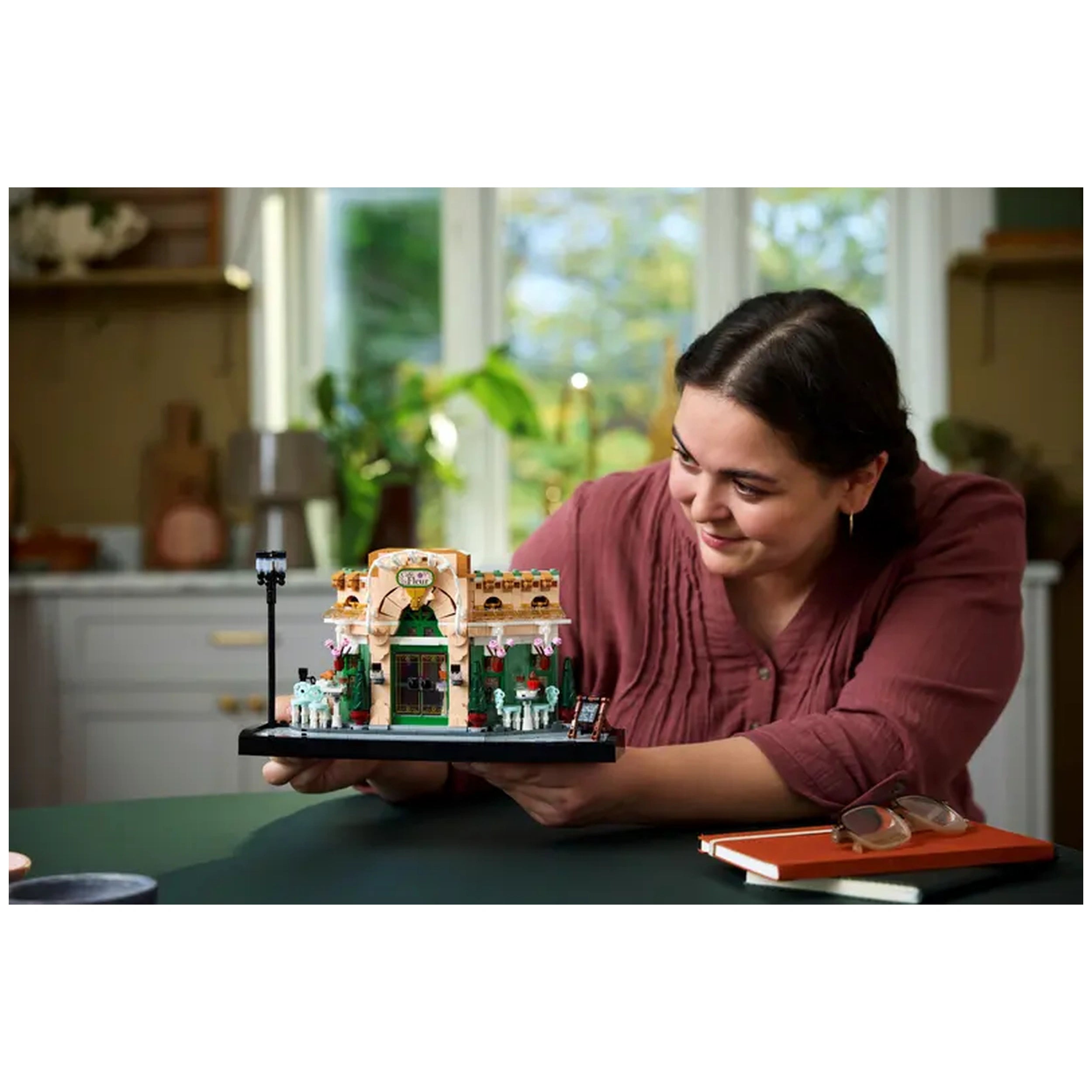 A woman smiles while holding the LEGO Icons French Café construction set, featuring vibrant details like awnings, flowerpots, and outdoor seating.