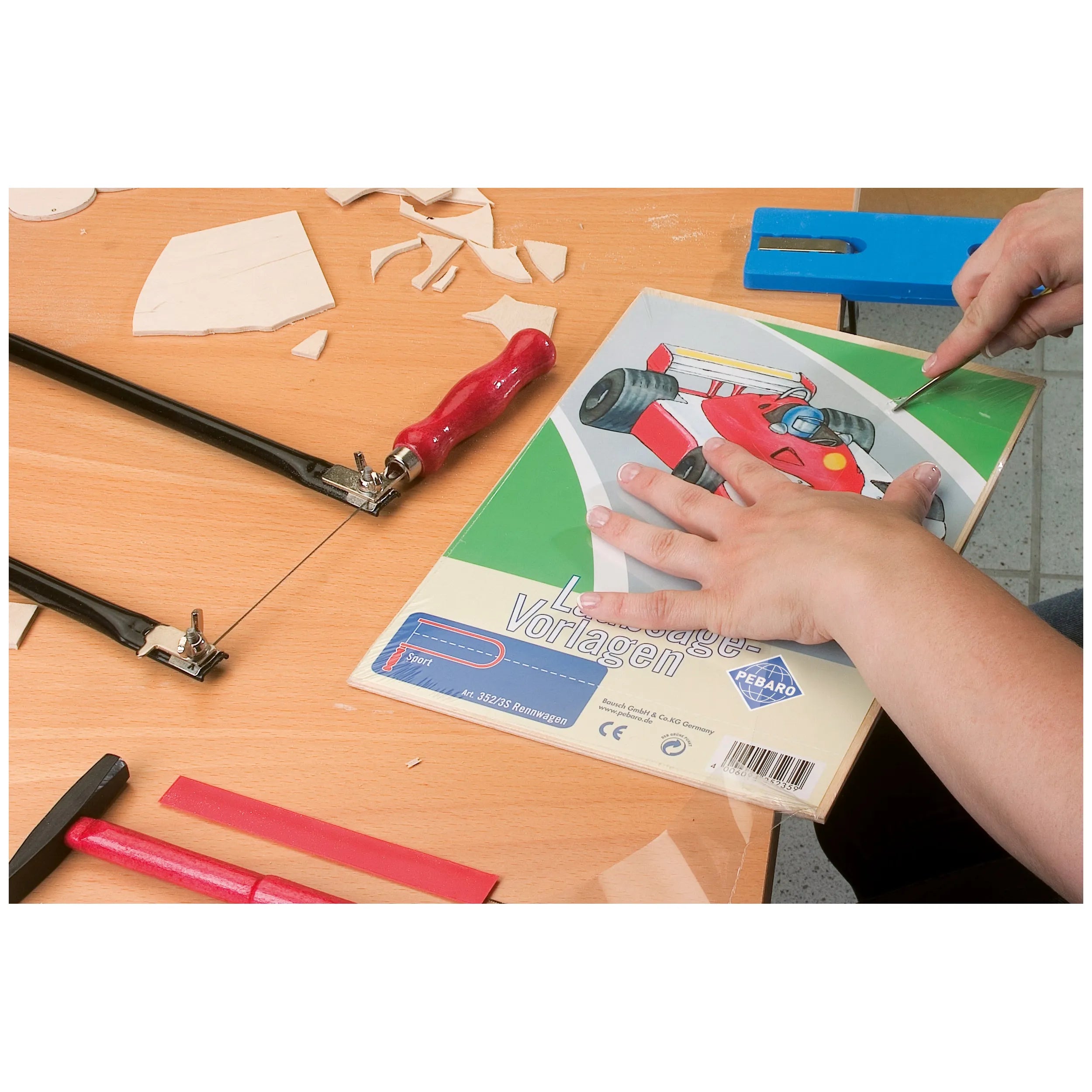 Hands assembling Prebaro Tractor model on birch plywood with fretsaws and craft tools on a wooden table.