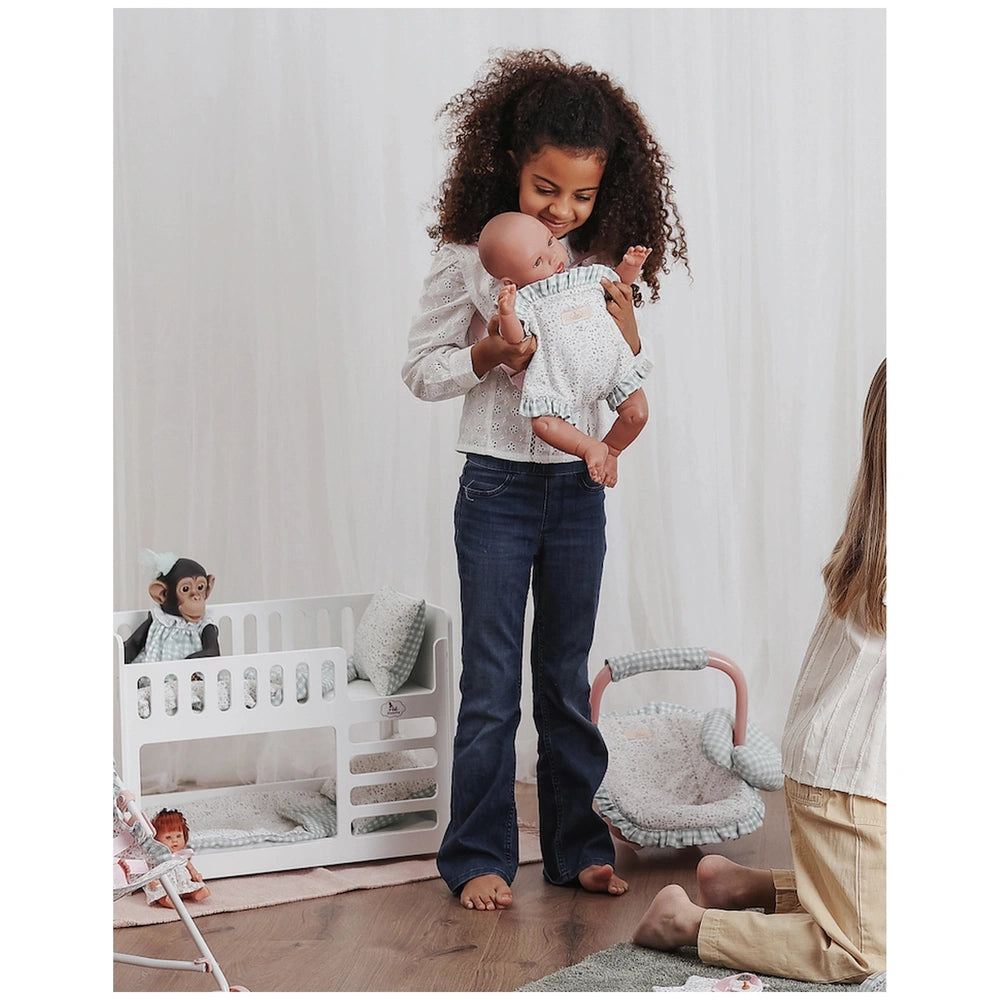 Child holding a doll in a baby carrier, smiling in a nursery setting with a crib and toys.
