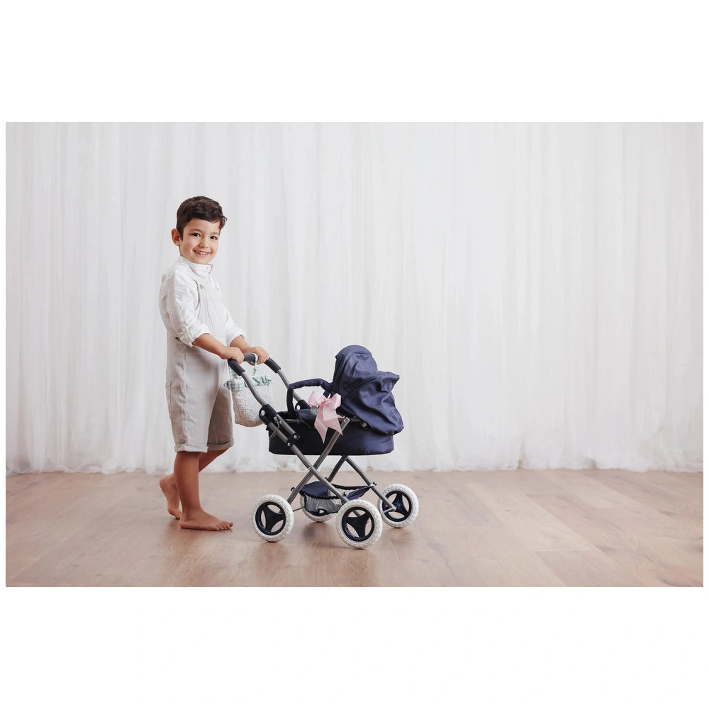 Child smiling while pushing a toy stroller with a navy blue hood, designed for ASI dolls, on a wooden floor in a bright room.