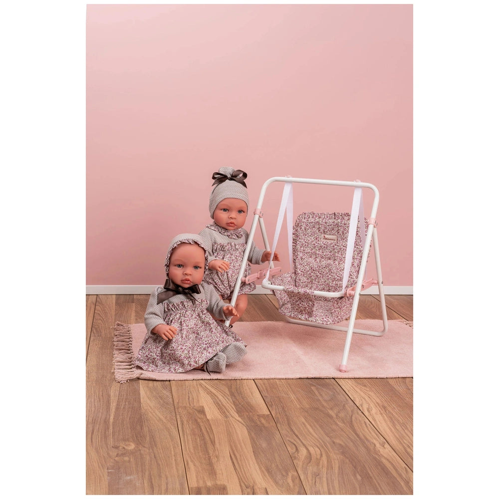 Two dolls in floral outfits sit next to a matching pink and white doll swing on a wooden floor.