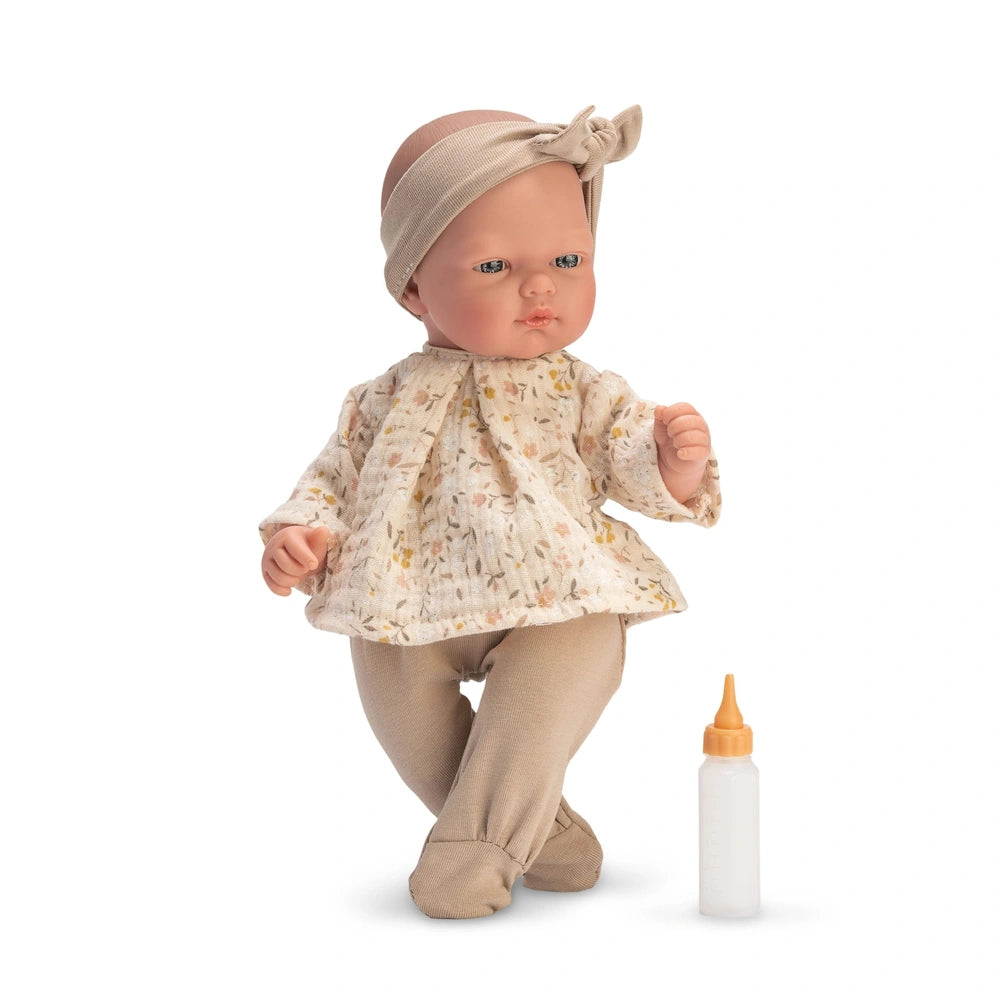 Bald doll with light eyes, beige headband, floral dress, beige leggings, standing by a toy bottle.