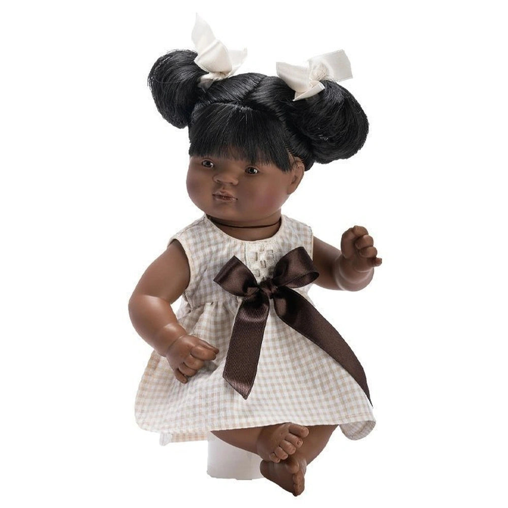 Samy 36 cm baby doll wearing a checkered dress with a brown bow, dual hair buns with white bows from the Sabana collection.
