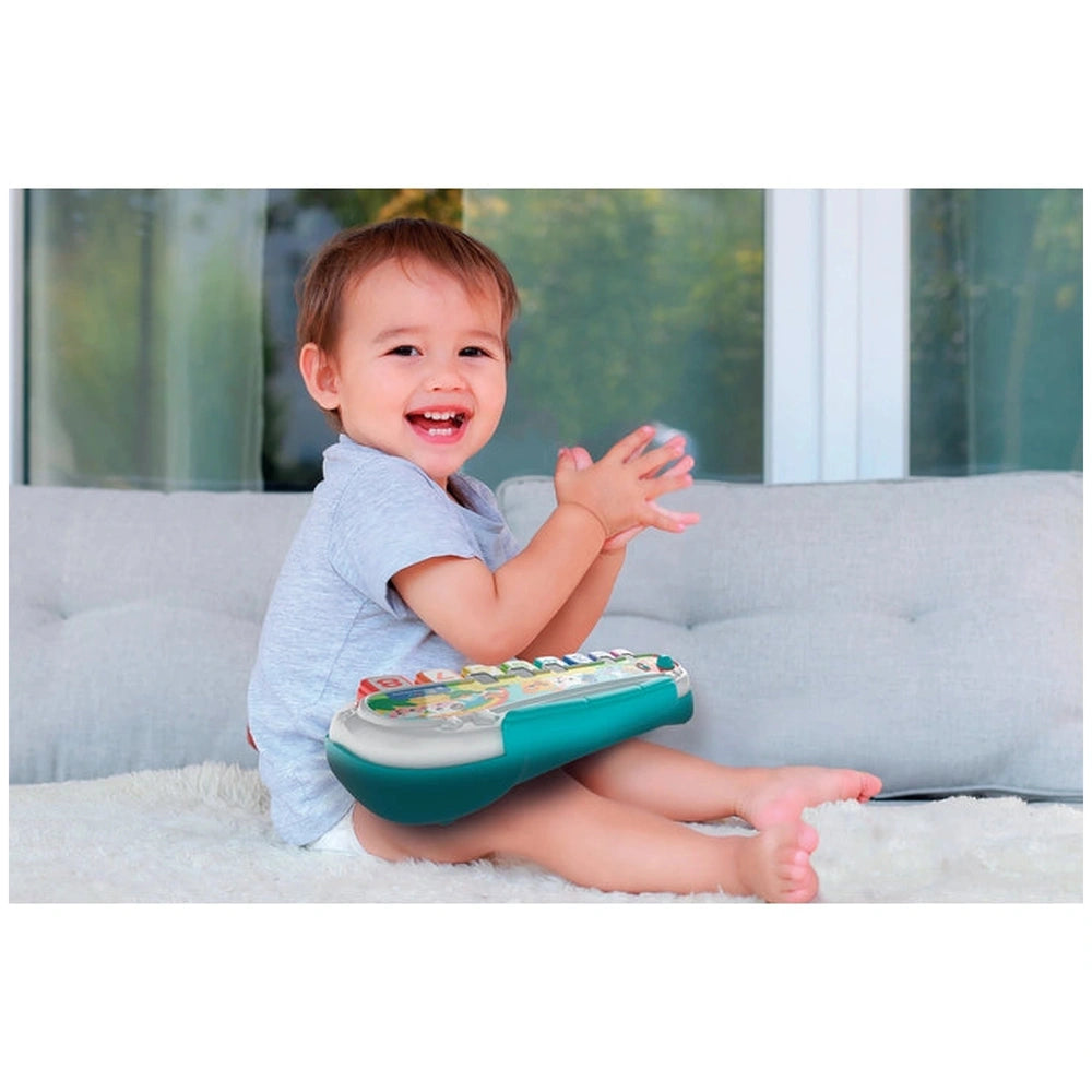 Baby Clementoni activity toy is held by a smiling child sitting on a soft surface, with a window view of greenery behind.