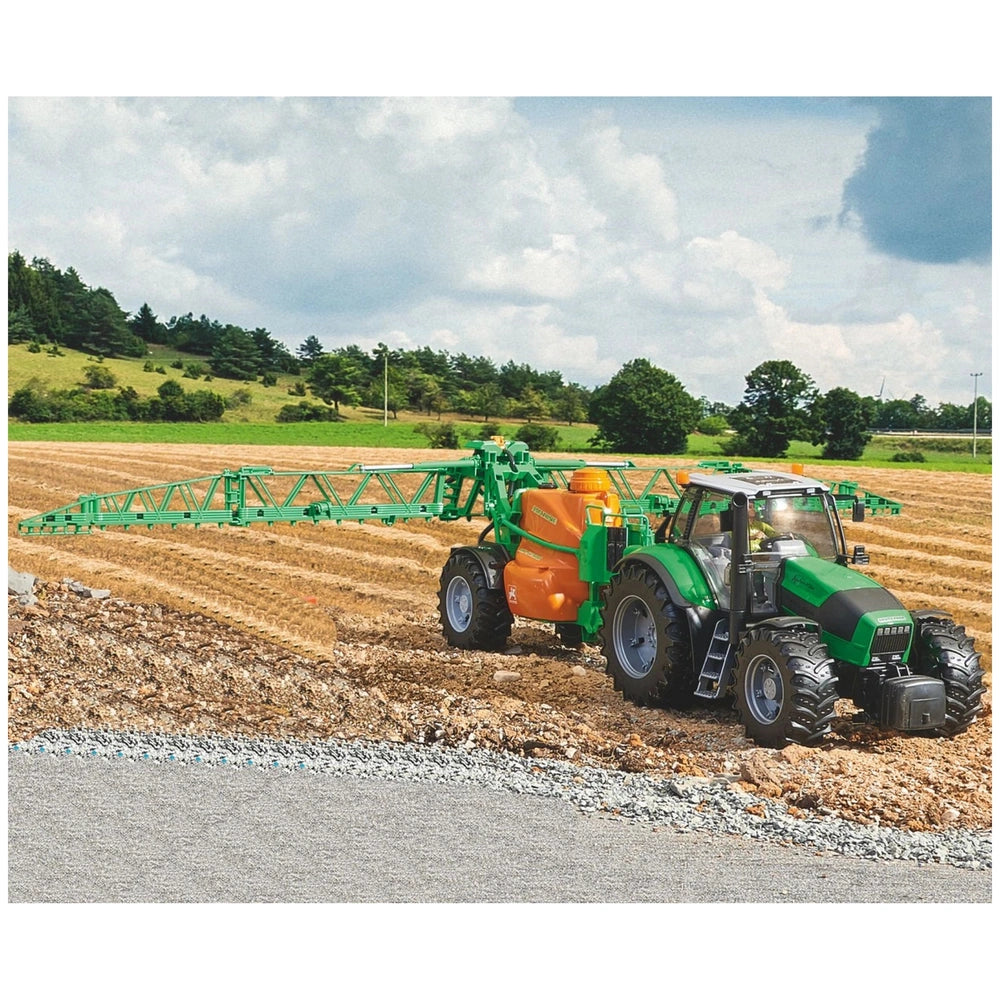 Toy tractor with green and orange field sprayer on a model farm landscape, showcasing realistic farming equipment.