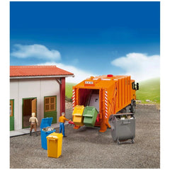 Toy garbage truck with colorful bins beside a small building, featuring miniature figures simulating waste collection.