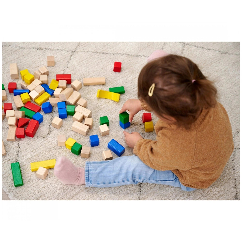 Eichhorn Baby Wooden Coloured Blocks are scattered around a child in a brown sweater and blue jeans, engaged in play on a car