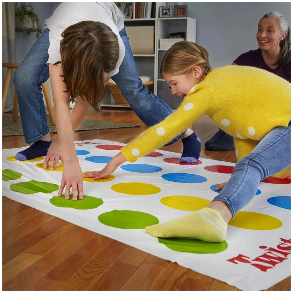 Twister Game board with colored circles, two children playing on a wooden floor, blue wall and white bookshelf in background.