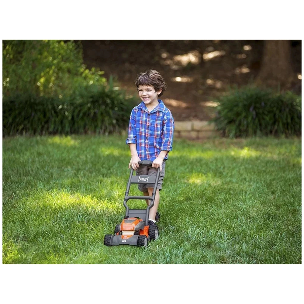Boy with brown hair in plaid shirt plays with black and orange Husqvarna toy lawn mower on green grass.