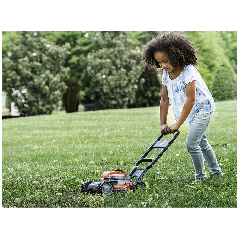 Girl with black hair in a blue shirt and pants pushes a black and orange Husqvarna toy lawn mower on green grass.