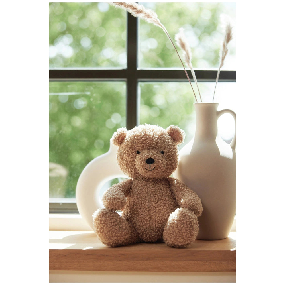 Brown teddy bear placed on a wooden surface with a white vase and green foliage visible through a glass window.