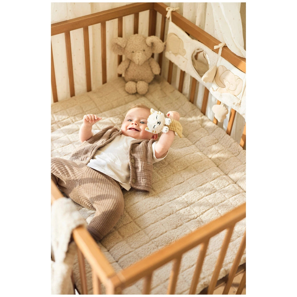 Jollein Wrist Rattle Tiny Park Charlie Cat rests beside a baby on a soft light brown crib mattress with wooden slats.