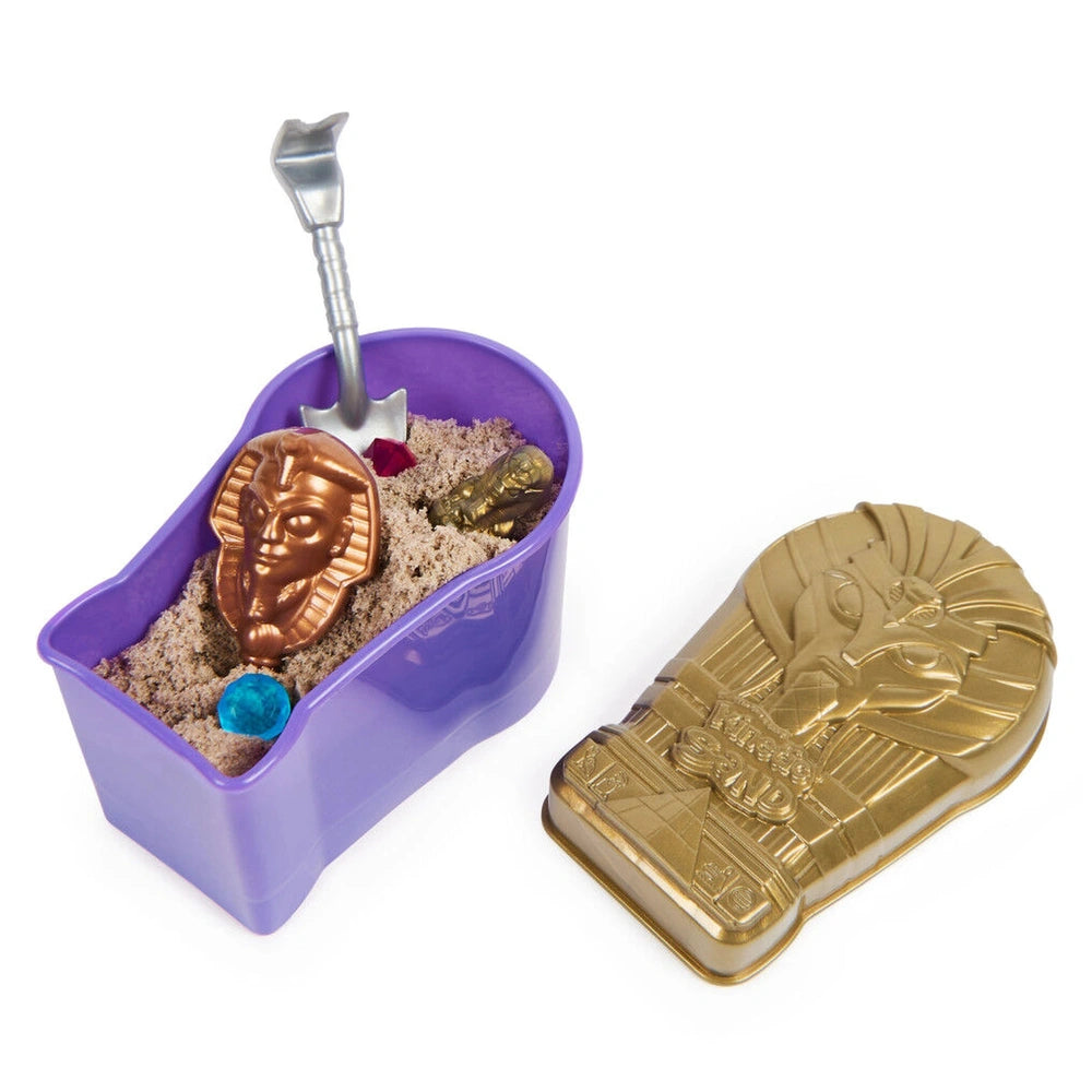 Kinetic Sand Mummy Tomb includes a purple container, light brown sand, a shiny golden object, a spoon tool, and a red flower.