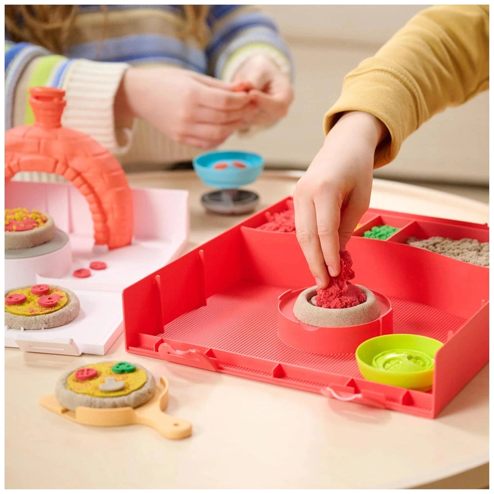 Kinetic Sand Squish Pizza toy is surrounded by colorful bowls and a red tray as a young girl plays at a table.