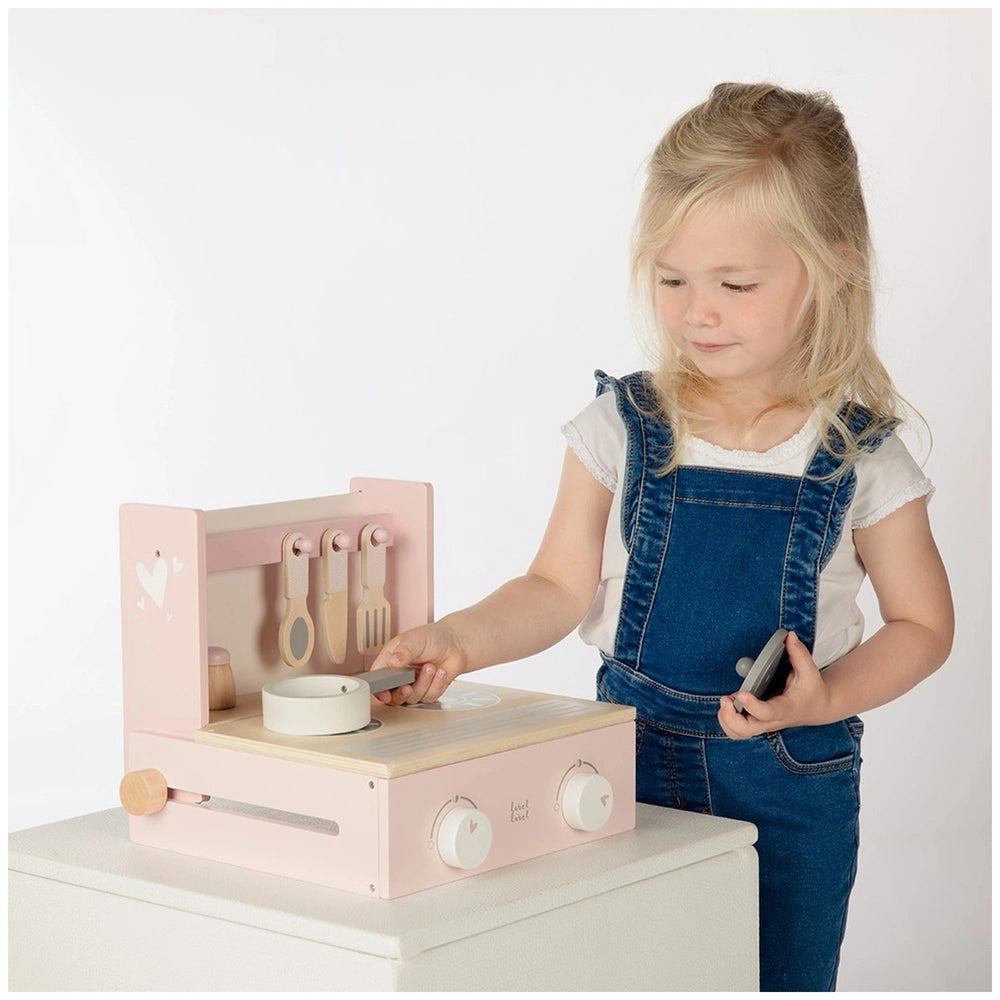 Foldable cooker toy kitchen in pink with white bowl, lid, wooden utensils, and a child wearing blue denim overalls.
