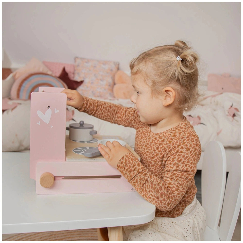 Child in leopard print top holds a pink heart-emblazoned pretend cooker at a white table, with pink and white patterned beddi