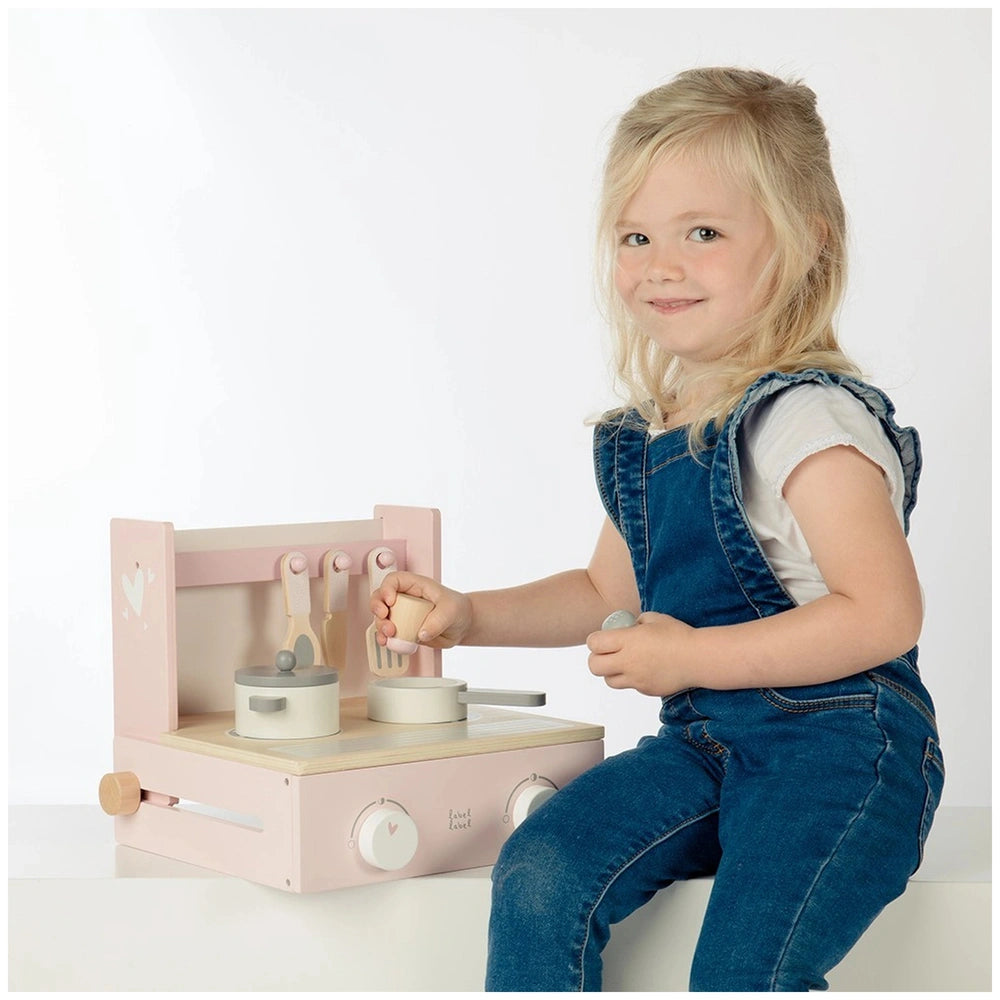 Foldable cooker toy in pink and white, with a young girl in a denim jumpsuit engaged in role-playing on a white tabletop.