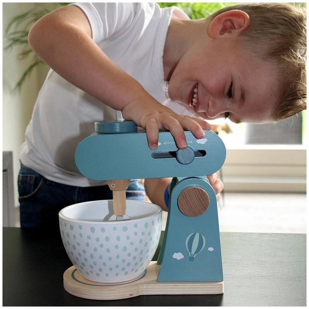 Child in white shirt and blue pants plays with a blue mixing device and white bowl with blue polka dots on a black table.