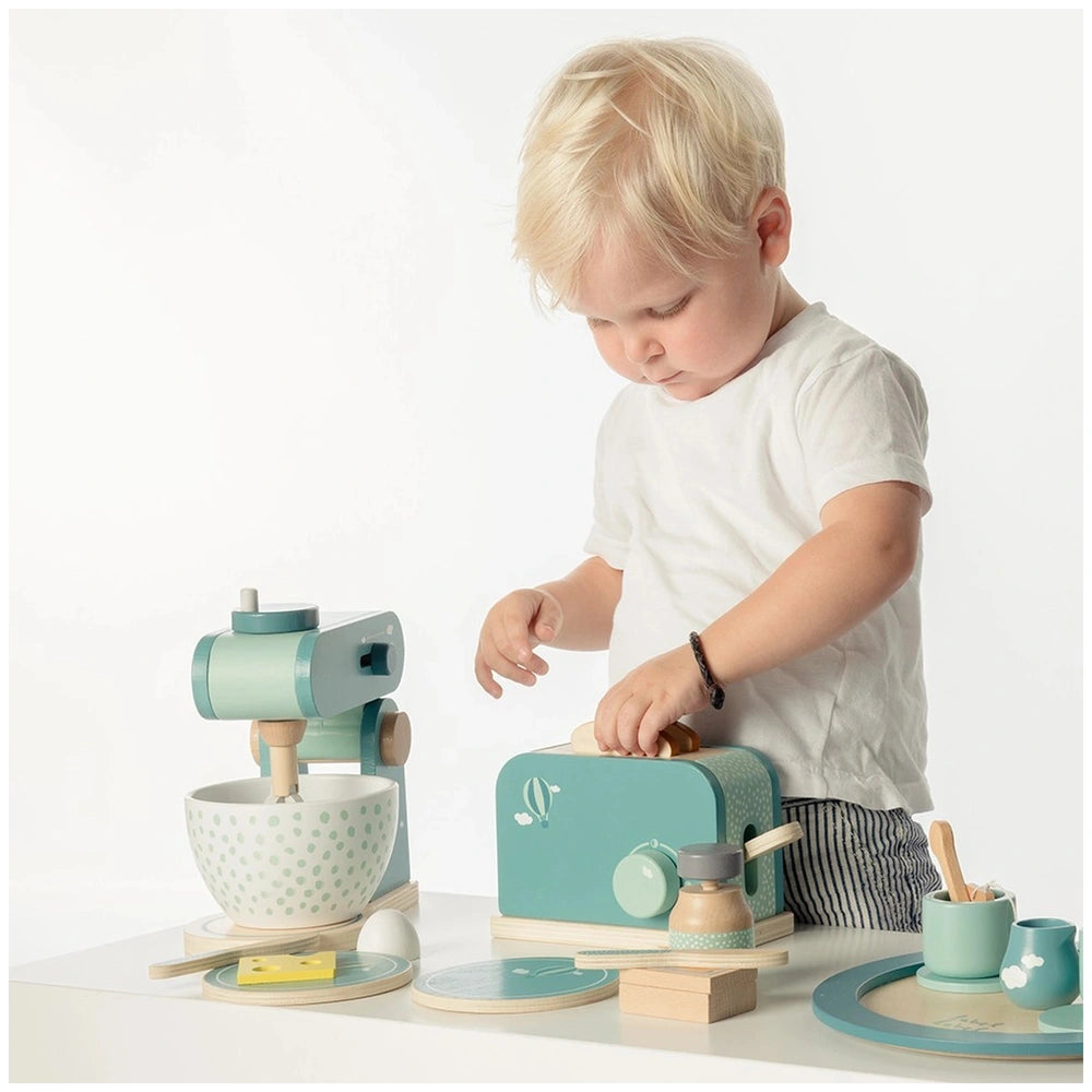 Child role-playing with a food processor set on a wooden table, surrounded by colorful plastic and wooden kitchen toys.