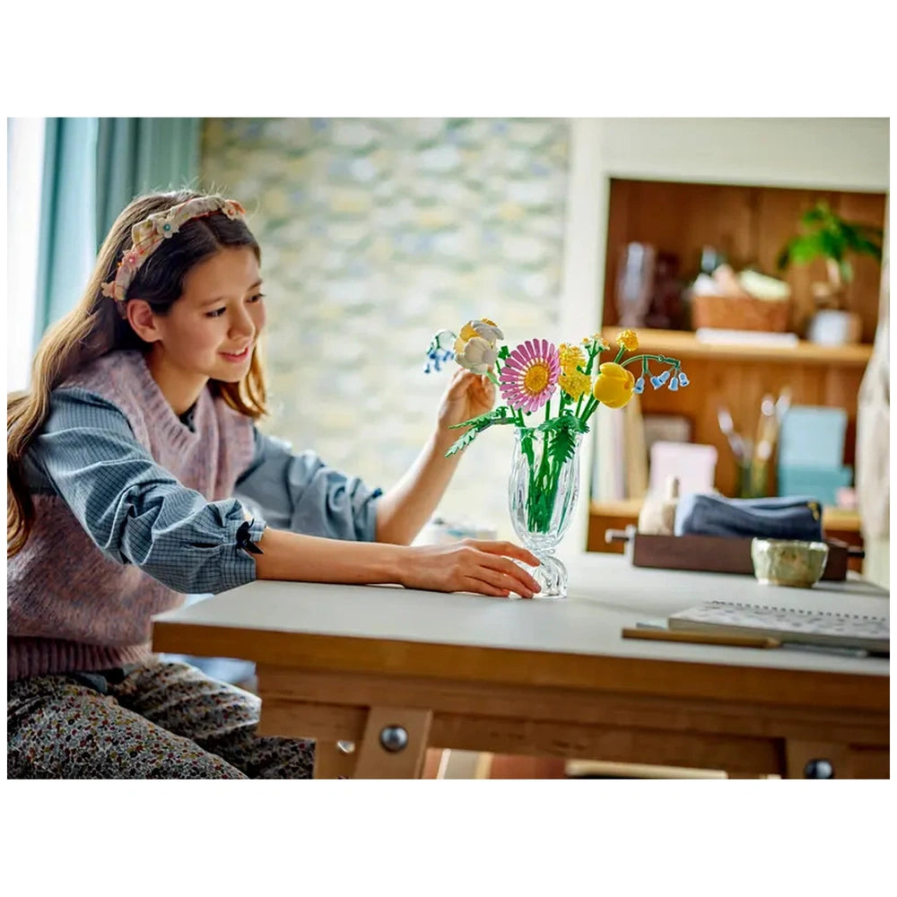 A young girl in a cozy room enjoys arranging a colorful LEGO® Botanicals Petite Sunny Bouquet in a clear vase on a wooden table.