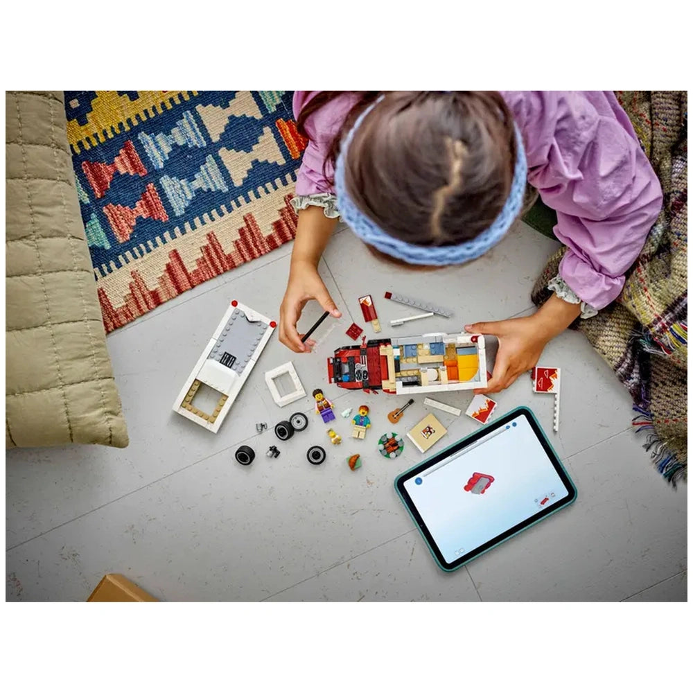 A child wearing a purple shirt and hairband assembles the LEGO® City Holiday Adventure Camper Van, surrounded by colorful bricks and accessories, with a tablet displaying building instructions nearby.