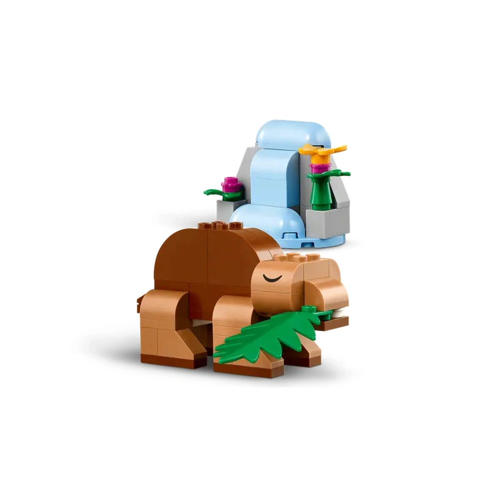 Brown LEGO dinosaur figure happily eating a green leaf, alongside a colorful brick-built plant and a chair with flowers.