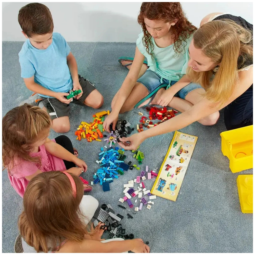 A group of four children and one adult sits on a gray carpet, surrounded by colorful LEGO bricks in various colors. They are sorting pieces, including blue, red, yellow, and green, while examining a LEGO instruction booklet.