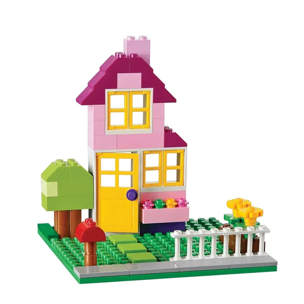 Colorful LEGO house with a pink and purple roof, yellow door, surrounded by greenery, a tree, and flowers in a playful layout.