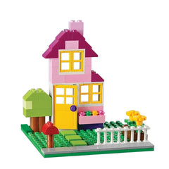 Colorful LEGO house with a pink and purple roof, yellow door, surrounded by greenery, a tree, and flowers in a playful layout.