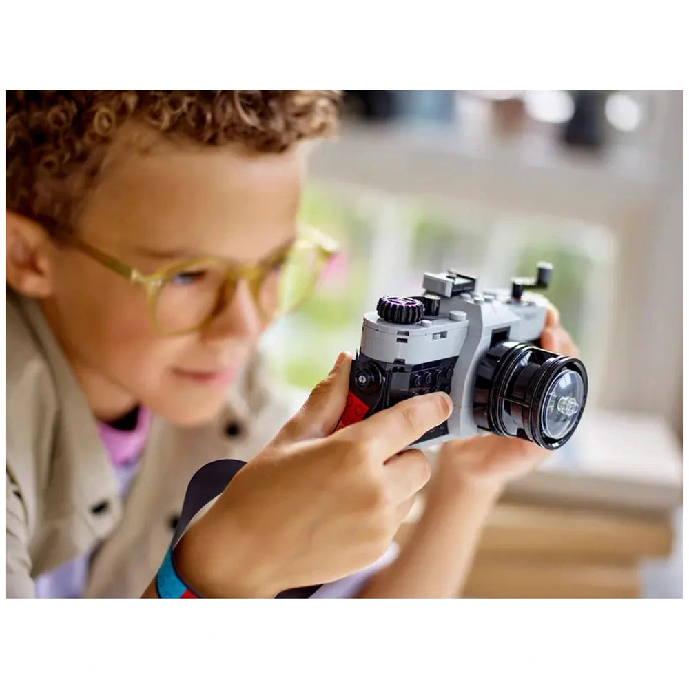 A young child with curly hair and yellow glasses holds a LEGO® Creator Retro Camera, focused on play. The camera features a sleek design with a black lens and moving parts, set against a bright background.