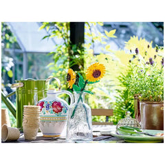 Two vibrant LEGO sunflower blooms in a clear bottle, surrounded by a decorative jug, green watering can, and potted plants, on a sunlit table.