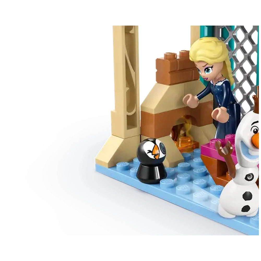 Elsa stands beside a fireplace in the Arendelle castle, excitedly interacting with a black and white penguin figure. Beside her, Olaf the snowman smiles cheerfully, amid colorful LEGO bricks and a cozy setting.