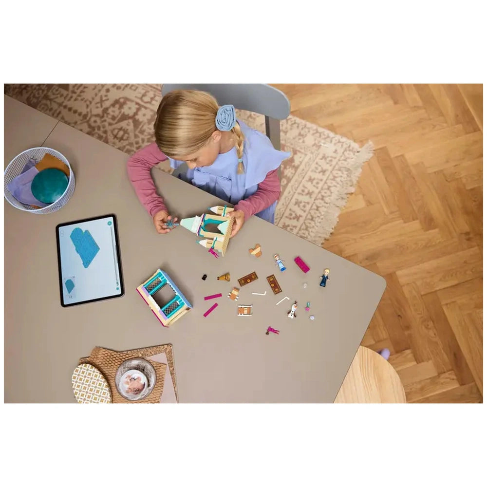 A child in a blue dress and pink sleeves focuses on building a LEGO Frozen castle, surrounded by colorful pieces and mini-doll figures on a beige table. An tablet displays instructions, with a cozy rug and wooden floor in the background.