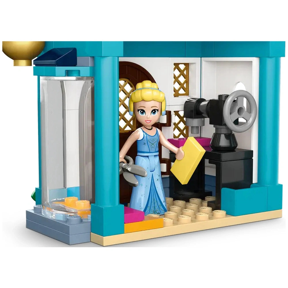 Cinderella, wearing a blue dress, holds a yellow block while standing in a colorful LEGO construction of a market. The scene includes a sewing machine, a charming door, and detailed walls, inviting imaginative play.