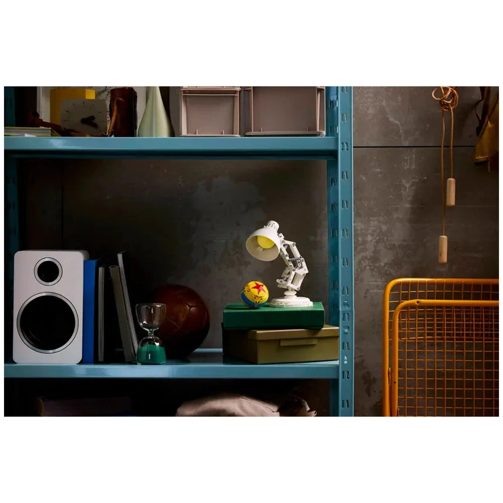 LEGO Luxo Jr. model on a green box, beside a starry ball, gray wall behind, with blue shelves, books, and a speaker.