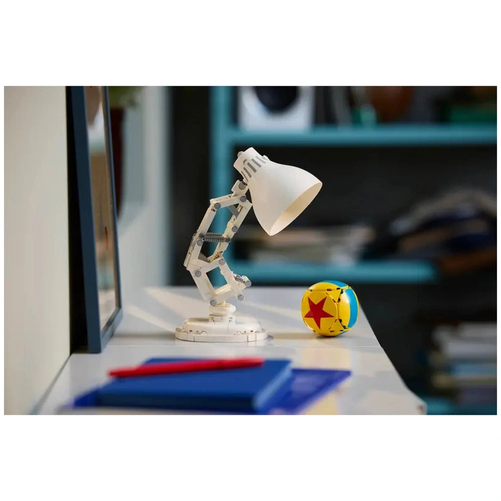 A white LEGO lamp with a flexible arm sits on a table next to a colorful ball featuring a red star, surrounded by blue notebooks and a red pen.