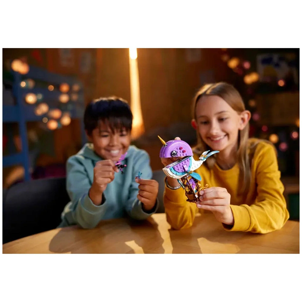Two children joyfully holding LEGO DREAMZzz Izzie's Narwhal Hot-Air Balloon, one displaying a tiny figure and the other the balloon, with a warm indoor setting.