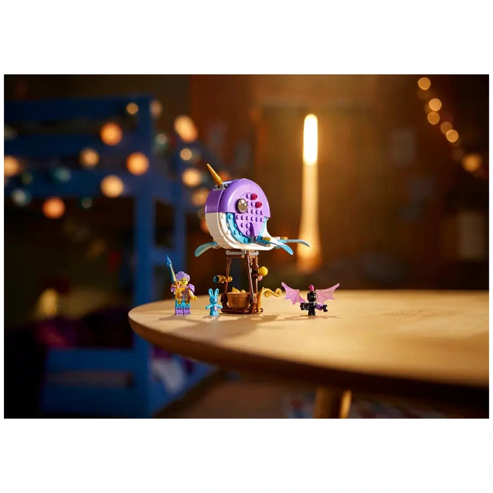 Colorful LEGO® DREAMZzz™ Izzie's Narwhal Hot-Air Balloon set on a wooden table, featuring a whimsical narwhal, mini figures, and a bunny. Soft lighting creates a cozy, imaginative atmosphere.