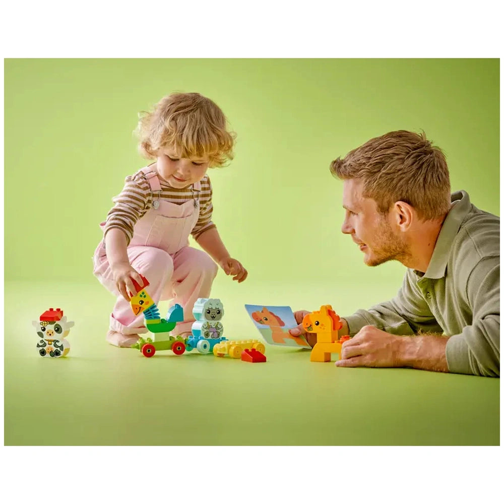 A young child in pink overalls plays with a colorful LEGO DUPLO Animal Train set alongside a smiling adult. Various buildable animal figures, including a rooster and a cow, are spread across a light green floor, with the adult engaging with a picture card.