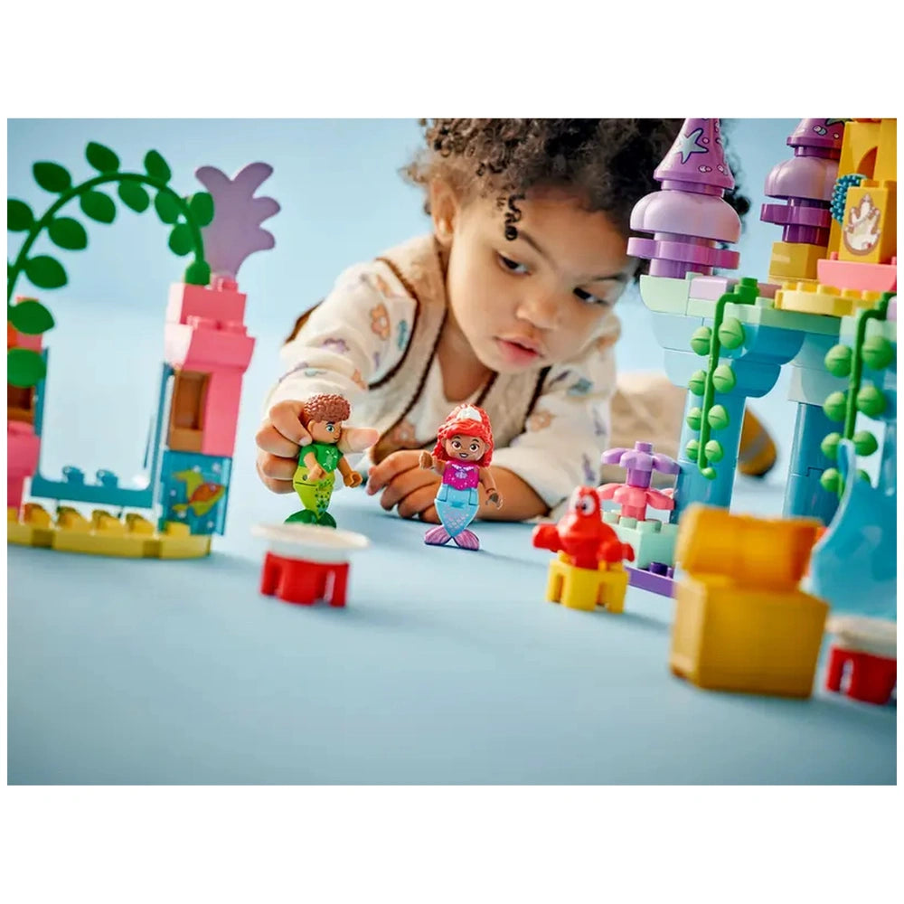 A young child plays on a blue mat, holding LEGO® DUPLO® figures of Ariel and a boy in front of Ariel's Magical Underwater Palace. Colorful building blocks and accessories surround them, creating a vibrant underwater scene.