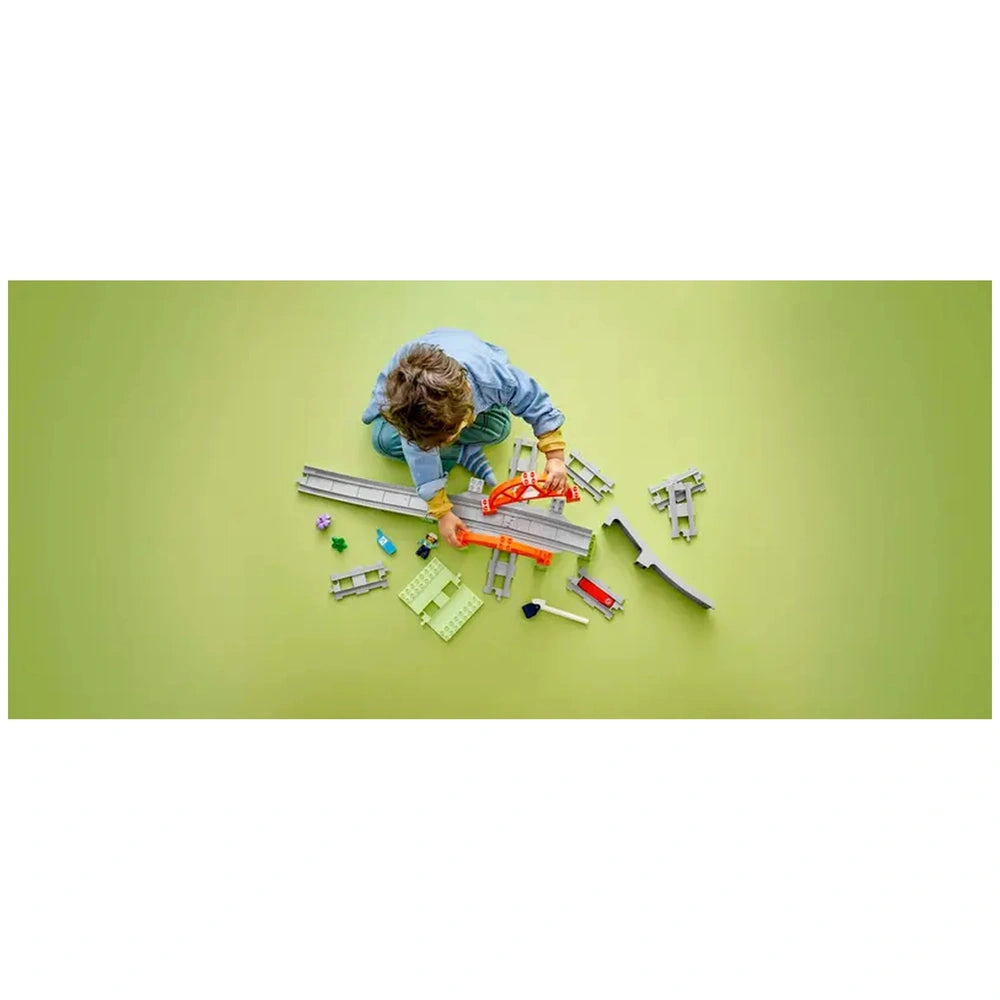 A young child sits on a green surface, building with the LEGO® DUPLO® Train Bridge and Tracks Expansion Set. Various colorful track pieces, a bridge, and small train vehicles spread around them, enhancing imaginative play and creativity.