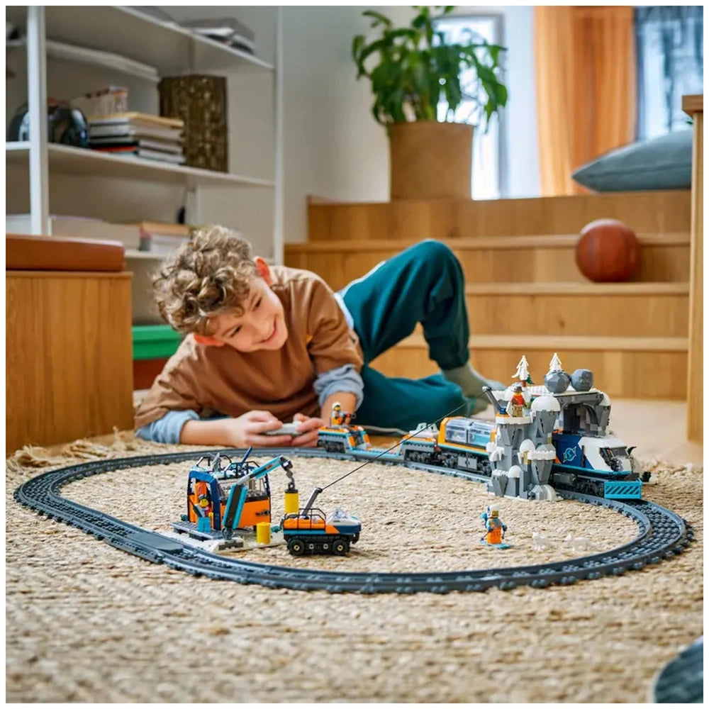 A cheerful child lies on a woven rug, controlling the LEGO® Explorers’ Arctic Polar Express Train set. The train winds around a snowy landscape with a mountain and toy figures, showcasing an engaging Arctic exploration scene.