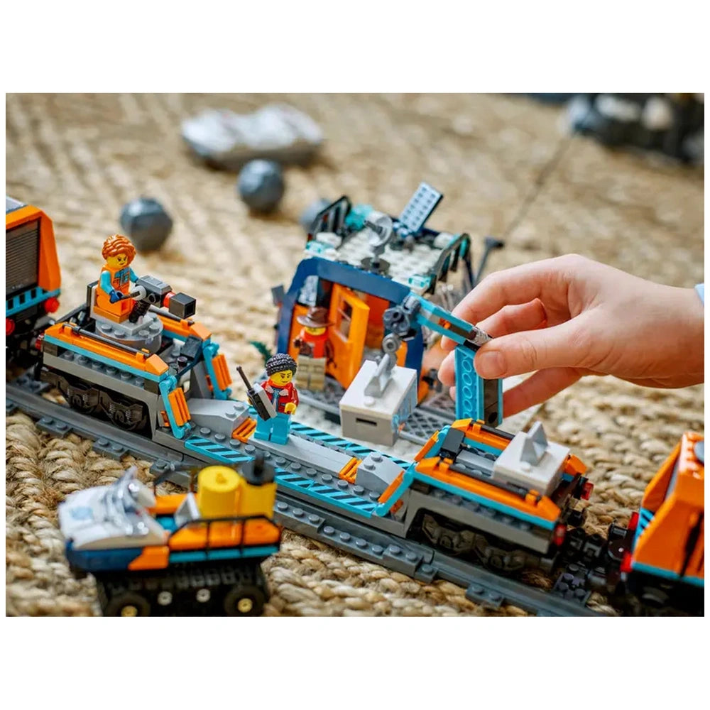 Colorful LEGO Arctic train set with two figures, a snowplow, and an outpost. A child's hand interacts with the build on woven texture.