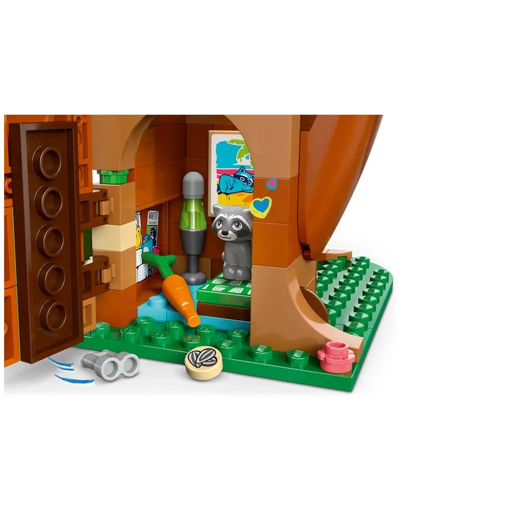 Inside a LEGO Friends tree house, a gray raccoon figure stands beside a vibrant green carrot on a grassy base. The brown door is slightly ajar, revealing colorful wall decor, including a picture with animals. A lamp and small items accompany the scene, inviting imaginative play.
