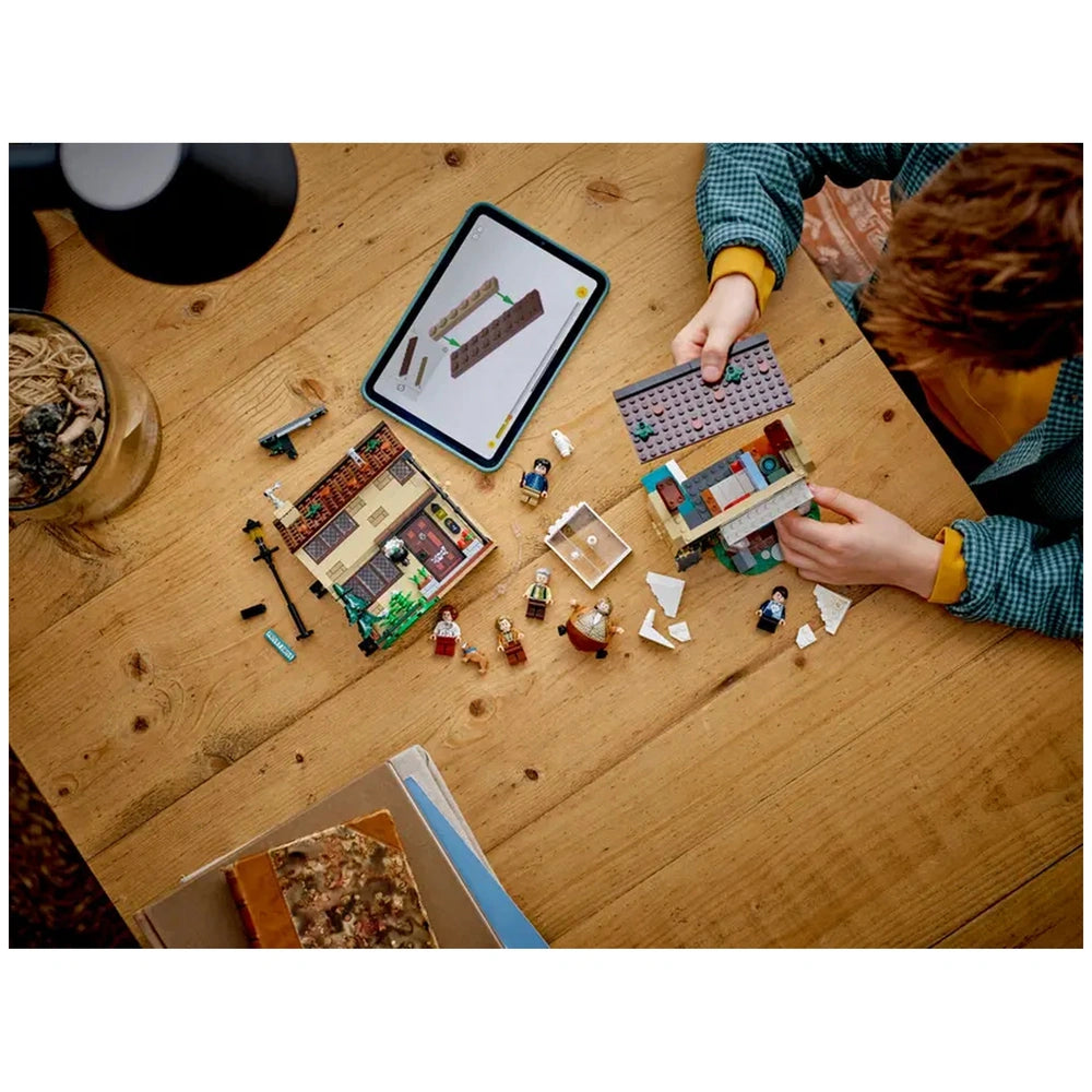 A child assembles the LEGO® Harry Potter™ Aunt Marge's Visit playset on a wooden table, surrounded by colorful bricks and mini-figures. A tablet displays building instructions nearby, with scattered pieces and a partially built house featuring a cozy interior.